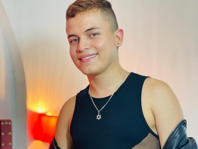 LeonAdams, young dude with short brown hair, smiling, wearing a black sleeveless top, silver star necklace, and black leather jacket, has a cute, slim, sexy build.