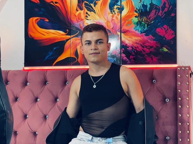LeonAdams, with short brown hair, wearing a black sheer tank top, black jacket off shoulders, white jeans, necklace, sitting on pink tufted bench, vibrant colorful wall art behind.