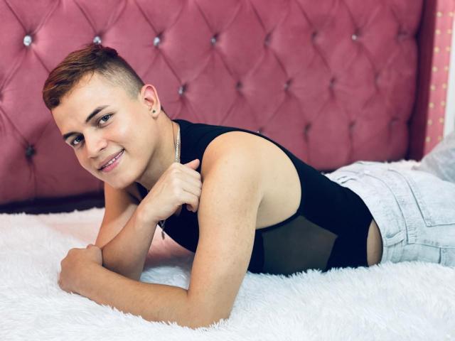 LeonAdams with short brown hair, black sleeveless top, and light jeans, lying on white fluffy bed, smiling, fingers near chin, pink tufted wall in background.