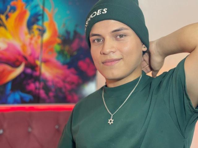 LeonAdams with a green beanie, green shirt, and star necklace, hand in hair, smiling. His medium brown skin and brown eyes are visible. He's wearing a green beanie.