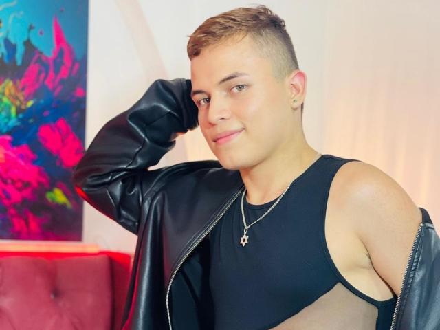 LeonAdams, a cute young dude with short brown hair, wearing a black leather jacket and sleeveless black top, showing off a cross necklace, with a playful, confident smile.