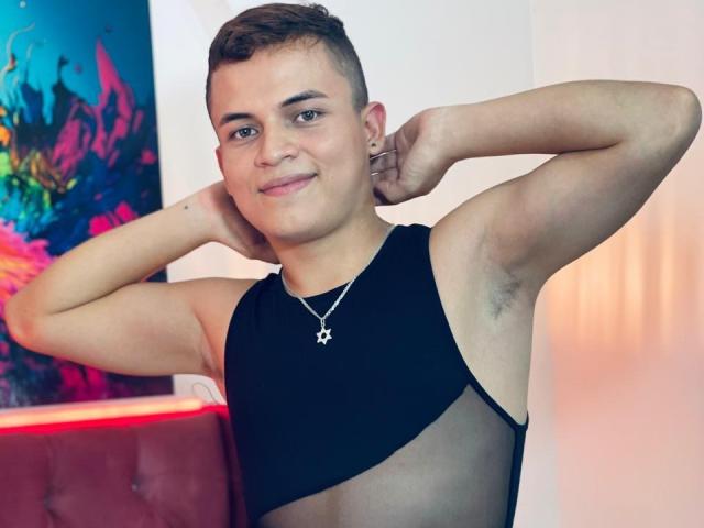 LeonAdams with short brown hair, smiling, arms raised, wearing a black sleeveless top with a star necklace, showing armpit hair. Visible chest and arms. Casual, confident.
