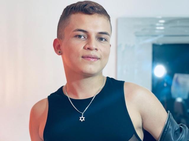 LeonAdams, short brown hair, sleeveless black top, silver star necklace, slight smile, visible ear piercing, leather jacket sleeve, light skin, blue eyes, slight muscle definition, casual pose.