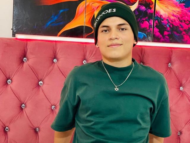 LeonAdams, wearing a green beanie and green shirt, with a silver necklace, has a cute face and a slight smile, sitting in front of a pink tufted couch.
