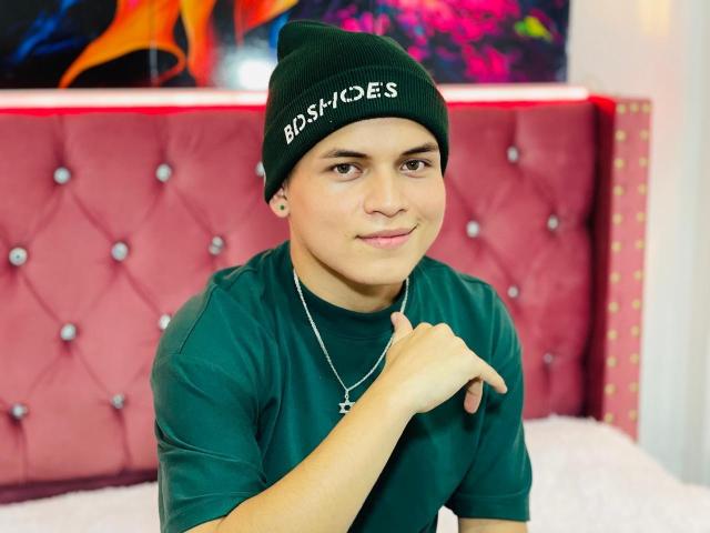 LeonAdams, wearing a green beanie and green shirt, with a silver cross necklace, has a cute face and is sitting with a subtle smile, holding his chin.