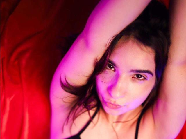 AphrilMercyy com os braços levantados, deitada em uma superfície vermelha, com iluminação roxa e rosa, olhos castanhos, lábios carnudos, cabelo preto, vestindo uma blusa preta com alças, olhando para cima..
