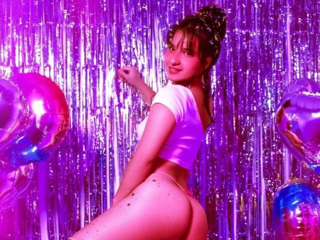 AphrilMercyy with her ass in a shiny purple top, looking back, her brown hair in a bun, surrounded by pink and blue balloons, in a purple-lit room.