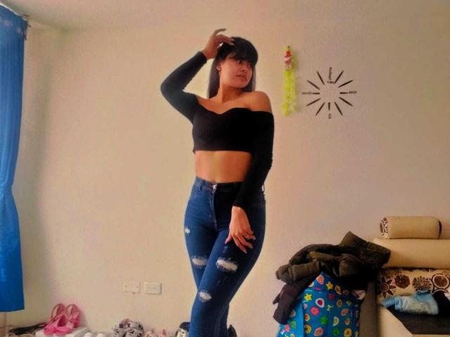 CrystallThonpsom with black off-shoulder top, blue ripped jeans, standing in room, hand in hair, medium tits, slim, dark hair, slightly smiling, casual, confident, jeans tight.