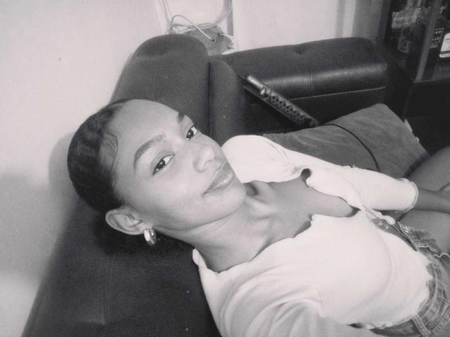 AylaCallowey with short braided hair, wearing a white cardigan and plaid skirt, lying on a dark couch, hand on chest, smiling, black and white photo, visible ear earring.