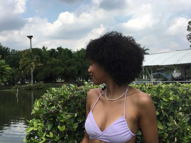 AylaCallowey with curly black hair, wearing a lilac halter top, showing her small tits and wearing a pearl necklace, standing by a green bush, looking to the side.