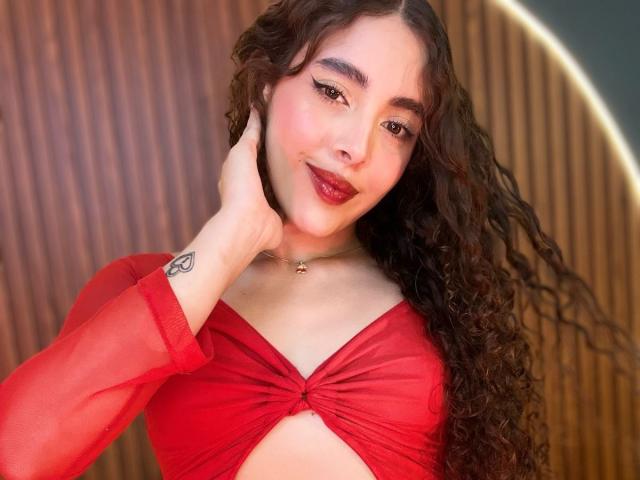 TianaVera with curly brown hair, red sheer top, small tits, tattoo on wrist, red lipstick, smiling, hand on cheek, wood background, glowing edge.