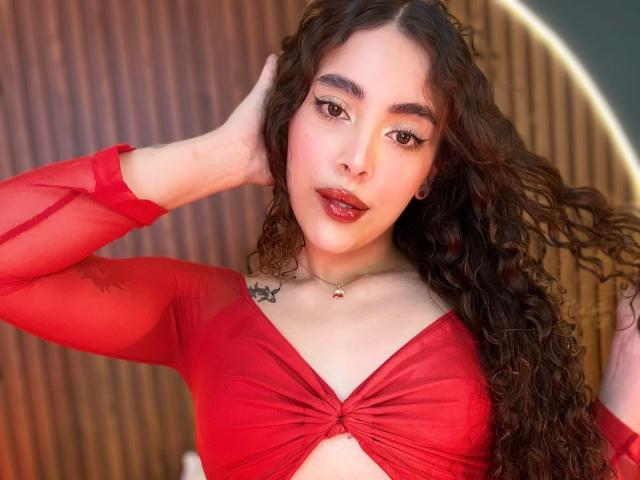 TianaVera with curly brown hair, red sheer long-sleeve top, tied-boob design, dark red lipstick, tattoo on left shoulder, hands in hair, wooden background, subtle makeup, confident expression.