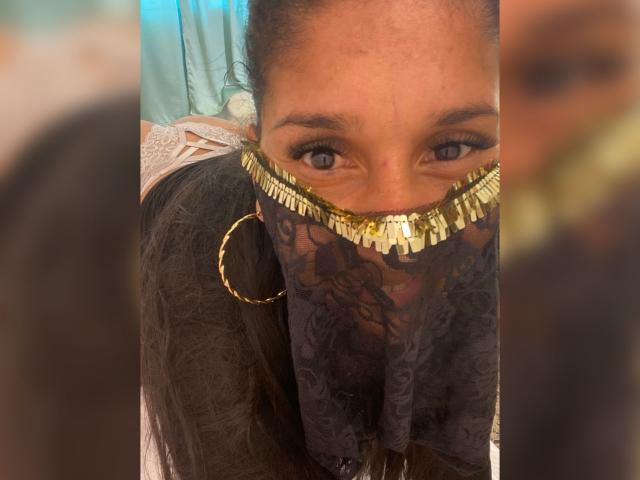 Le visage de KiraTaleb, partiellement dissimulé par un masque en dentelle noire orné de franges dorées, révèle ses yeux bleus. Elle a les cheveux foncés et porte des boucles d'oreilles dorées..