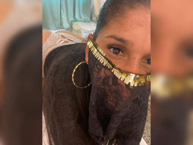 Kira Taleb, met donker haar, draagt een zwart kantmasker met gouden rand, grote gouden oorringen en witte kanten lingerie, en kijkt in een spiegel..