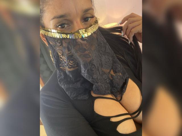 KiraTaleb usa uma máscara de renda escura com detalhes dourados, um top preto com recortes que mostra seios grandes, segura o cabelo, tem olhos castanhos olhando para cima, cabelo preto longo e unhas pretas..