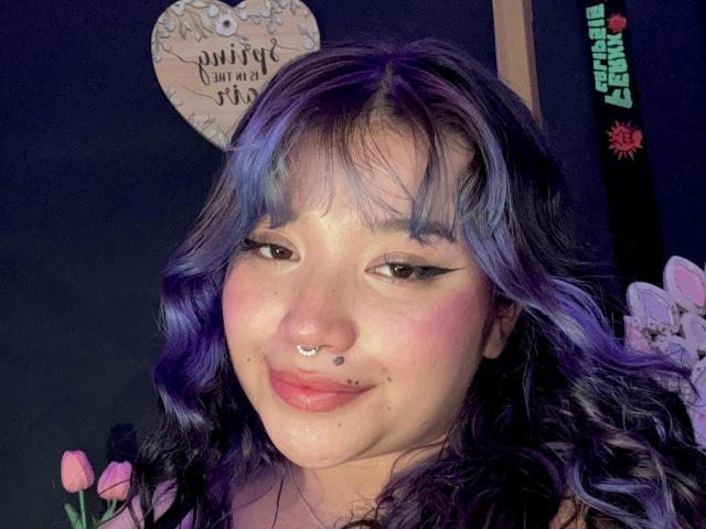 HonnyBlaice, com cabelo roxo-azul, piercing no nariz e no lábio, sorrindo com delineador preto, blush rosa e sardas, mostrando um rosto fofo, muito fofo..