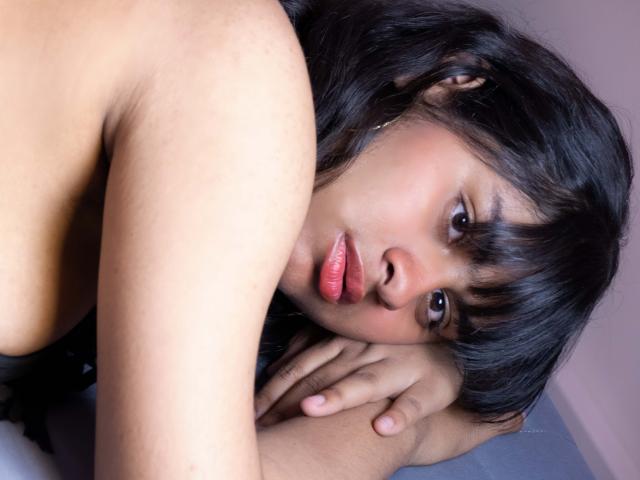AnanyaSyed with dark hair, pouty pink lips, and brown eyes, leaning on her hand, topless, showing her shoulder and looking at the camera with a slight frown.