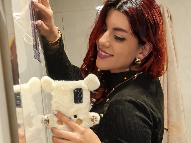 ZoeeMar mit roten Haaren, schwarzem Oberteil, hält eine weiße, flauschige Teddybär-Handyhülle, lächelt, trägt goldene Ohrringe, schaut in den Spiegel, die rechte Hand ist erhoben, die linke Hand hält den Teddybären..