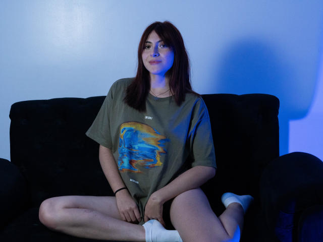 SpencerLe, met rood haar, zit op een zwarte bank, gekleed in een oversized groen T-shirt, witte sokken en geen broek, met gespreide benen, waarbij haar vagina zichtbaar is. Blauw licht accentueert haar..