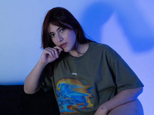 SpencerLe with dark brown hair, green t-shirt featuring a colorful wave design, and a contemplative expression, touching her chin with a blue-lit background. Her ass is partially visible.