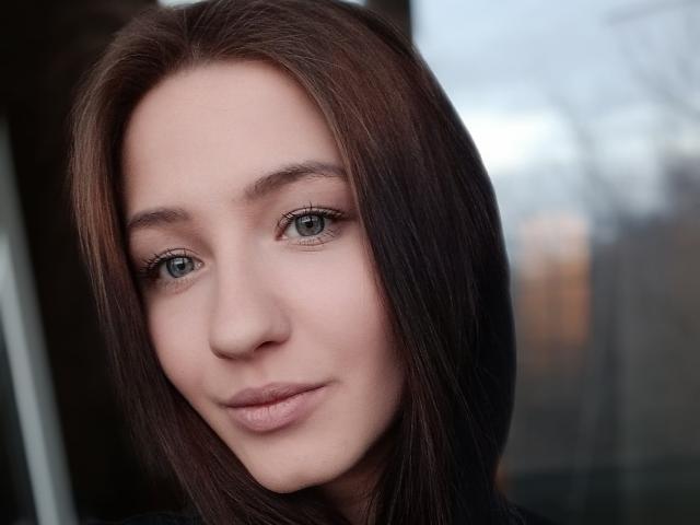 Le visage de RoxieSara, avec ses yeux vert clair, son rouge à lèvres rose discret et ses cheveux bruns lisses, arbore un sourire léger et confiant. Sa peau est douce, son maquillage est naturel et son expression est calme et attrayante..