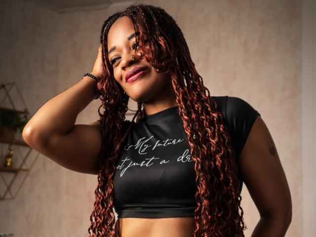 LucyCastillo with curly red braids, black crop top, smiling, hand in hair, showing her tits, medium brown skin, pink lipstick, in a well-lit room.