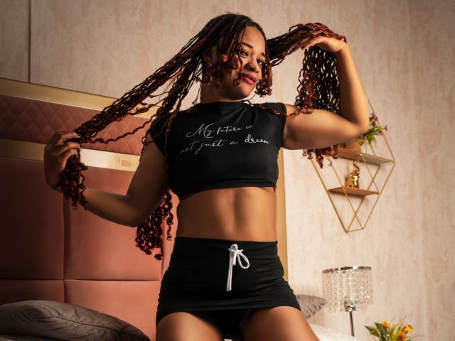 LucyCastillo with curly brown braids, smiling, wearing a black crop top and mini skirt, holding her hair, showcasing her toned midriff and small ass. Bedroom setting, warm lighting.