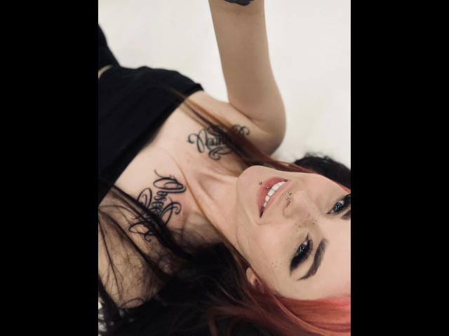 Coco1996-hot with red hair, black eyeliner, nose ring, smiling, black top, tattoos on chest, lying down, holding phone, black arms, white background, visible breasts, no pants.