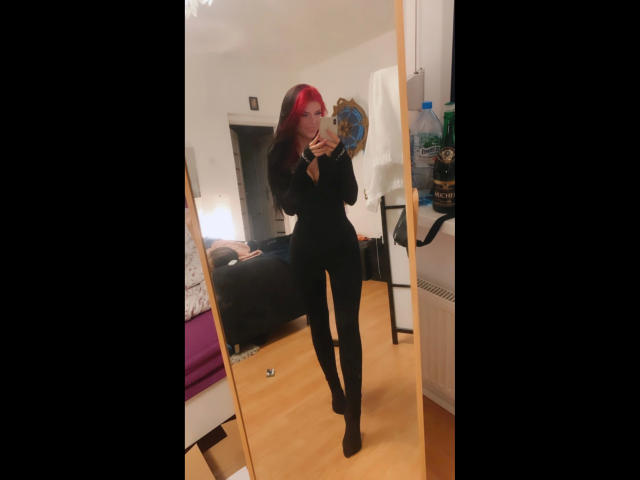 Coco1996, avec ses cheveux roux, sa tenue noire et ses chaussettes noires, prend un selfie devant un miroir, montrant ses fesses fines et ses petites seins, tout en tenant un téléphone dans une chambre..