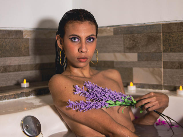 ElevenSkys, with wet dark hair, holding purple lavender, in a bathtub, looking at camera, wearing gold hoop earrings, water covering her small tits and ass, candles in background.