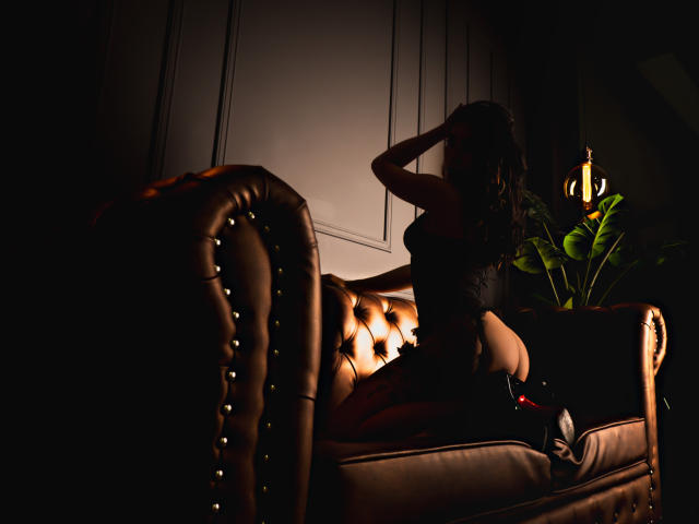 AnyaJay with wavy brown hair, dark lingerie, and black heels, kneeling on a tufted brown leather couch, hands in hair, under warm light, with a potted plant nearby.