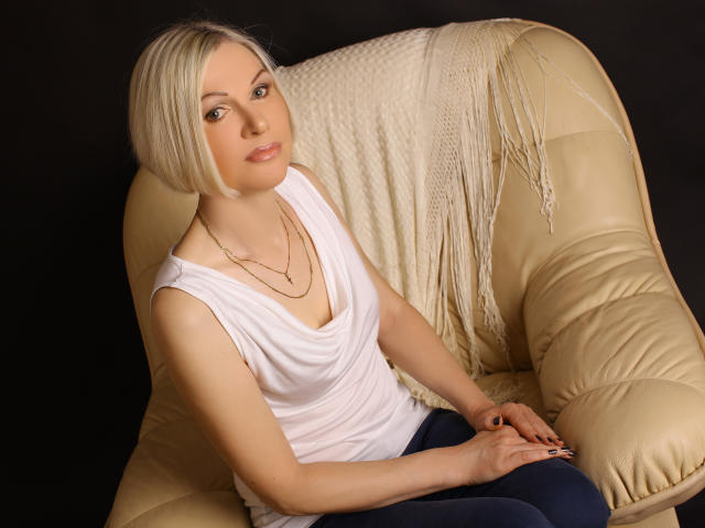CityGlam has blonde bob, wearing white sleeveless top, blue pants, gold necklace, sitting on beige leather chair with white knitted blanket, looking at camera with subtle smile.