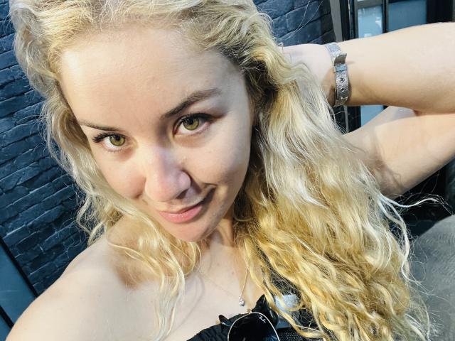 CamiDallx with wavy blonde hair, green eyes, and a subtle smile, wearing a black top and silver bracelet, looking up at the camera. Her ass is partially visible.