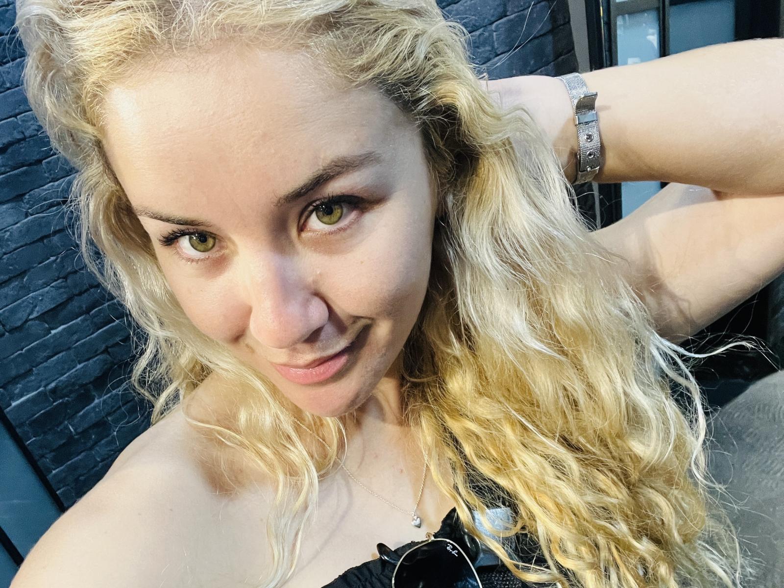 CamiDallx, con cabello rubio ondulado, ojos verdes y una sonrisa sutil, vestida con una blusa negra y una pulsera de plata, mirando hacia la cámara. Su trasero es parcialmente visible..