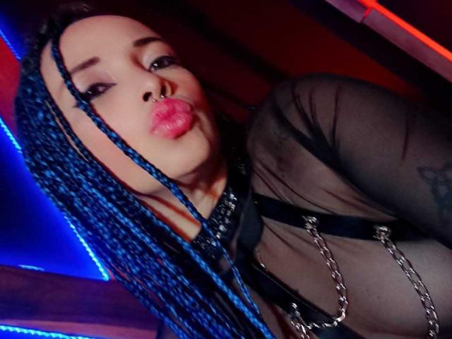 DalilaMusa with blue braids, nose ring, and pink lips, pouting, wearing a sheer black top with metal chains, showing her tits, looking sexy.