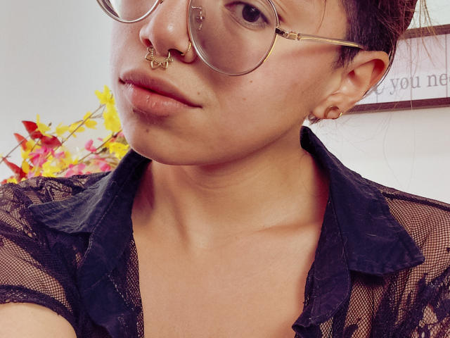 NaturalMariaHun, usando óculos, um piercing no nariz e brincos, veste uma blusa preta de renda, tem uma expressão neutra e cabelo curto e escuro. Seus seios são pequenos e sua vulva não está visível..