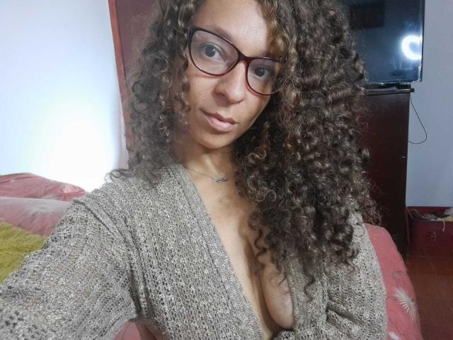 Angelonix, con i capelli castano ricci, indossa occhiali e un cardigan grigio a maglia, mostrando il seno con un profondo scollo a V, seduta su un divano rosa..