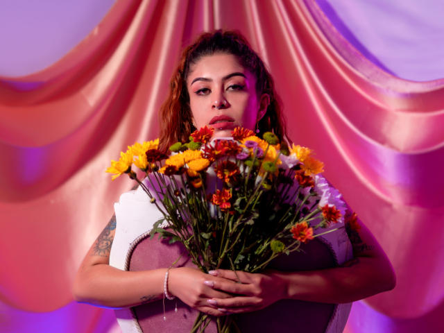 AlaiaLove holding a bouquet of yellow and orange flowers, with tattoos on her arm, wearing a white and purple top, pink and purple lighting.