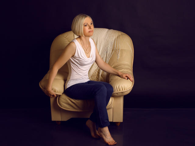 CityGlam met kort blond kapsel, een witte tanktop en blauwe jeans zit zonder schoenen op een beige fauteuil, met armen wijd, en kijkt naar de zijkant. Er ligt een crème deken op de stoel..