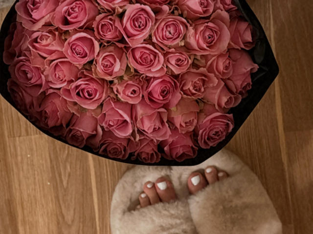 SugarrX's feet, covered in fluffy white socks, stand below a black-wrapped bouquet of pink roses on a wooden floor. Her toenails are painted white.