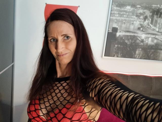 NadjaNeah-hot with long dark brown hair, blue eyes, wearing a black fishnet top showing her large, red, mesh-covered tits. She's smiling, standing in a room.