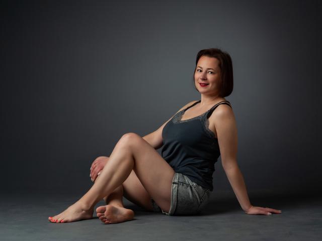MarieSho sits on the floor, legs crossed, wearing a dark tank top and denim shorts, her red-painted toenails visible, with a relaxed, smiling expression.