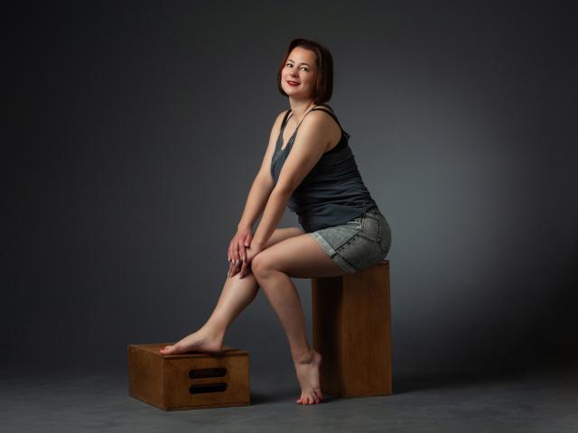 MarieSho, barefoot, sitting on a wooden block, wearing a black tank top and denim shorts, legs crossed, smiling, red lipstick, short brown hair, dark gray background.