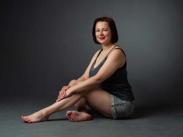 MarieSho met kort bruin haar, rode lippenstift, een grijze tanktop en denim shorts, zittend met gekruiste benen, lachend, met blote voeten, rode nagellak op haar tenen, en in een ontspannen houding..