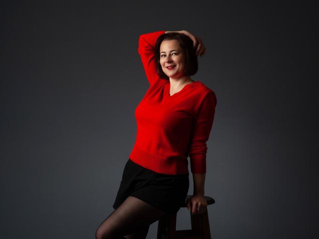 MarieSho with red sweater, black skirt, and black stockings, sitting on stool, hand on head, smiling, showcasing her tits and ass. Dark background, studio light.