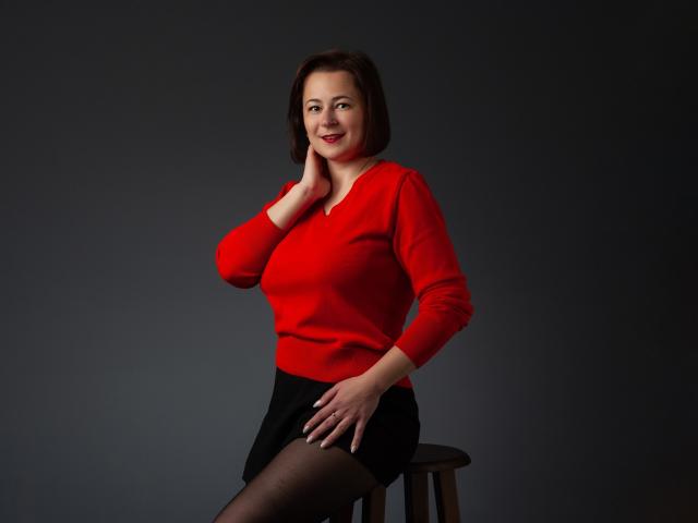 MarieSho with short brown hair, red sweater, black skirt, and black stockings, sitting on a stool, hand on neck, other hand on knee, smiling at the camera.