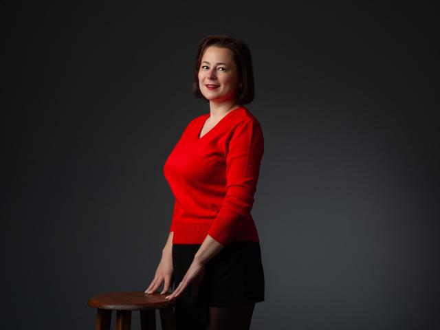 MarieSho with short brown hair, red sweater, black skirt, standing, hands on wooden stool, smiling, medium tits, dark background, studio shot.