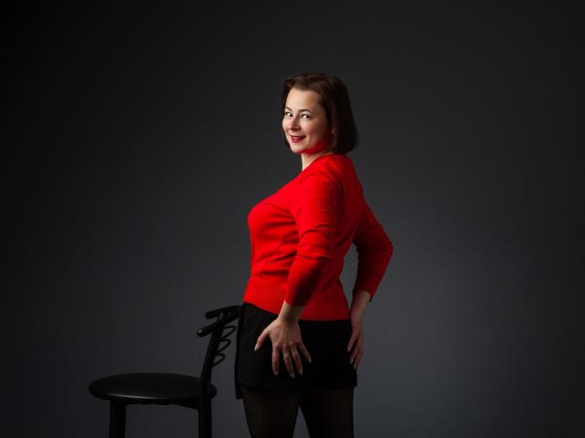MarieSho stands with hands on hips, wearing a red sweater and black skirt, next to a black chair, showing her ass and a cute smile.