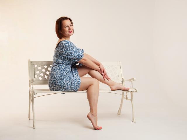 MarieSho with short brown hair, blue floral dress, sitting on a white metal bench, legs crossed, barefoot, smiling at camera, right arm on bench, left arm on knee.