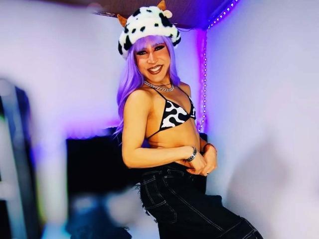 DarianaGio with purple hair, cow-pattern bikini top, black skirt, and cow hat, smiling, showing small tits, in a room with purple LED lights.